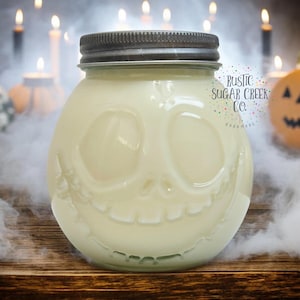 Peut inclure: Une bougie blanche dans un pot en verre en forme de crâne avec un sourire narquois. Le pot a un couvercle argenté et le texte "Rustic Sugar Creek Co. Handmade" est imprimé sur le côté.