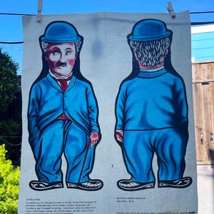 May include: A fabric panel featuring a blue and white illustration of a Chaplin-esque figure in a blue suit and bowler hat. The figure is shown from the front and back. The panel is hanging outside on a clothesline.