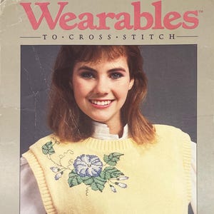 Può includere: Una donna indossa un gilet giallo a punto croce con un motivo floreale. Il gilet ha uno scollo tondo ed è sovrapposto a una camicia bianca con colletto. L'immagine ha il testo "Wearables" e "To Cross Stitch" in rosa.