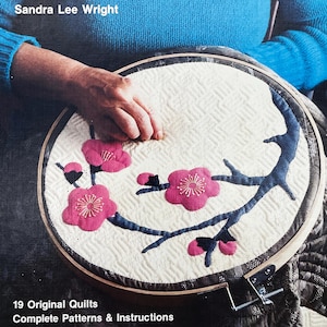 May include: A quilted embroidery hoop featuring a floral design with pink blossoms and blue branches. The text "19 Original Quilts Complete Patterns & Instructions" is visible at the bottom. The artist's name, "Sandra Lee Wright", is at the top.