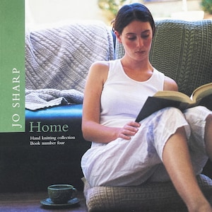May include: A woman reading a book, seated on a floor cushion. The book's title is not visible. The cover of the book is black. The text "Home" is visible, along with the author's name "Jo Sharp" and the words "Hand knitting collection Book number four".