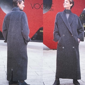 May include: A charcoal grey, floor-length, double-breasted coat with a wide collar, long sleeves, and a back vent. The garment features a button-front closure and is displayed in a Vogue Patterns advertisement with the text VOGUE PATTERNS.