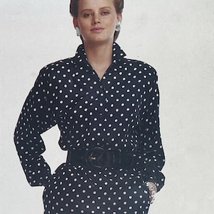 May include: A black dress with white polka dots, featuring a collared neckline and long sleeves. The dress is cinched at the waist with a wide black belt. The model is wearing large white earrings and a bracelet.