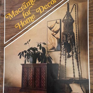 May include: A vintage book cover titled Macramé for Home Decor featuring a brown macramé plant hanger with twisted cords, a dark wooden cabinet with a herringbone pattern, a potted green plant, and a framed abstract wall art piece.