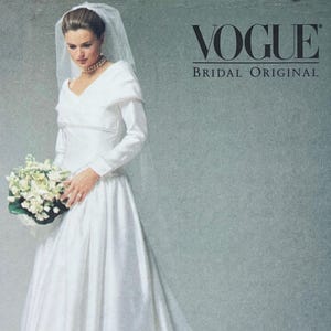 May include: A white bridal gown with long sleeves and a V-neckline. The dress features a full skirt and is adorned with delicate beading. The bride is holding a bouquet of white flowers and wearing a veil. The Vogue Bridal Original logo is visible.