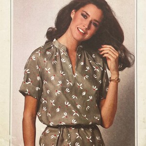 May include: A short-sleeved olive green blouse featuring a repeating pattern of small white petals with red centres. The garment has a notched neckline, a stand-up collar, and cuffed sleeves, paired with a matching belt at the waistline.