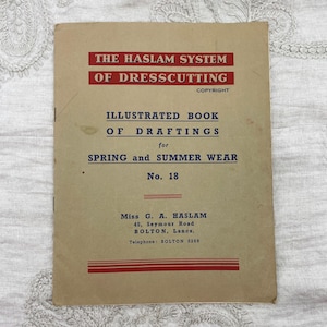 Op de afbeelding: Een beige papieren boekje getiteld The Haslam System of Dresscutting, Illustrated Book of Draftings for Spring and Summer Wear, No. 18. De omslag bevat rode en blauwe tekst, inclusief de auteur Miss G. A. Haslam en adresgegevens.