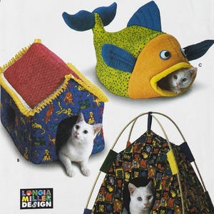 Puede incluir: Tres camas para gatos diferentes, una casa, un pez y una tienda de campaña. Cada cama está hecha de tela colorida con un patrón divertido. La casa es roja y azul con un borde amarillo. El pez es amarillo y verde con aletas azules. La tienda de campaña es marrón y amarilla con un patrón blanco y negro. Cada cama tiene un gato blanco dentro.