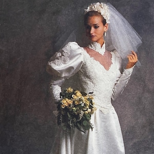 Peut inclure: Une robe de mariée blanche avec des manches longues, un col montant et un corsage transparent. La robe présente des détails de dentelle complexes et des manches bouffantes. La mariée tient un bouquet de fleurs jaunes et porte un voile et une coiffe florale.