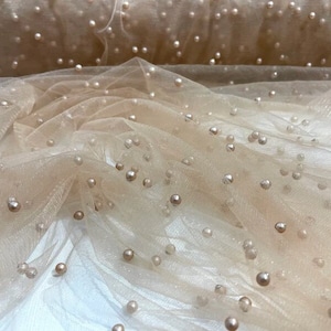 May include: Beige tulle fabric with scattered pearl-like embellishments. The fabric has a soft, delicate texture and a subtle shimmer.