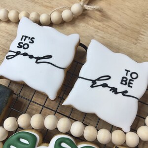 以下が含まれることがあります： 黒いアイシングで飾られた白いシュガー クッキーが 2 つ。1 つのクッキーは枕の形をしていて、黒い線が引かれた「It's so good」と書かれています。もう 1 つのクッキーは枕の形をしていて、黒い線が引かれた「To be home」と書かれています。