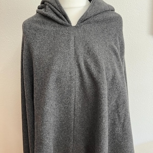 May include: A gray hooded poncho displayed on a mannequin. The poncho is made of a soft, textured fabric and features a hood and a split front. The bottom hem has a unique, uneven cut. The garment is a solid, neutral gray color.