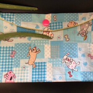 May include: A blue fabric pouch with a patchwork design featuring cartoon dogs and bones. The pouch has a yellow zipper and a pink button.