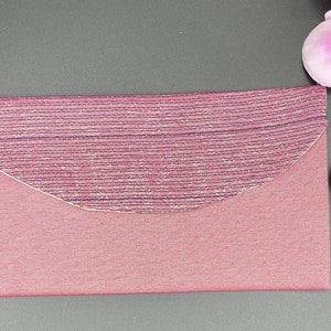 Puede incluir: Un sobre de tela rosa con una solapa decorativa, texturizada y plateada. La solapa tiene un borde curvo y un brillo sutil.
