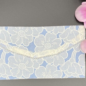 Op de afbeelding: Een lichtblauwe bloemenclutch met witte bloempatronen. De clutch heeft een witte rand en een zilveren kralenketting sluiting. De achtergrond is een donkergrijze ondergrond met roze orchideeën.