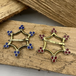 Könnte beinhalten: Zwei verzierte goldfarbene Gürtelschnallen mit sternförmigem Design. Eine Schnalle hat blaue Emaille-Akzente und die andere rote Emaille-Akzente.