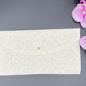 Op de afbeelding: Een witte stoffen envelop met een bloemenpatroon en een gouden knoopsluiting.