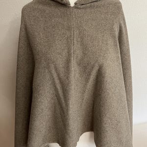 May include: A hooded cape in a neutral taupe color. The cape features a hood and a flowing, asymmetrical hemline. The fabric appears to be a soft, wool-like material. This garment is designed for warmth and style.