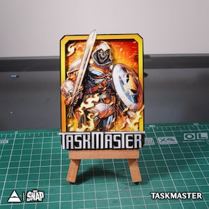 May include: A cardboard cutout of Taskmaster, a Marvel Comics superhero, holding a sword and shield. The cutout is on a small wooden easel and has a yellow background with flames.