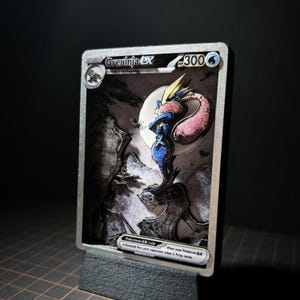 Może przedstawiać: Kolekcjonerska karta 3D typu shadow box przedstawiająca Greninja EX z 300 HP. Karta ukazuje niebiesko-różową postać stojącą na skalistym klifie na tle dużego białego księżyca. Tekst zawiera napisy Greninja EX, Stage 2 oraz Pokemon EX rule.