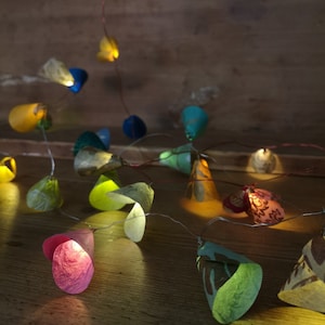 Könnte beinhalten: Eine Lichterkette mit bunten Papier-Lampenschirmen. Die Lichter sind auf einem Kupferdraht aufgefädelt und leuchten, wodurch ein warmes Licht entsteht. Die Lampenschirme sind in verschiedenen Farben und Mustern gehalten, darunter Grün, Blau, Gelb, Rosa und Rot.