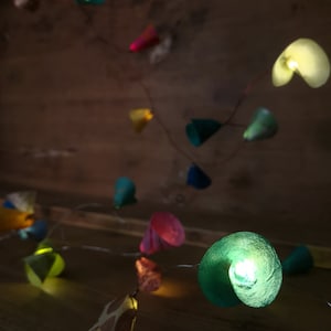 Puede incluir: Una cadena de luces de papel de colores con bombillas LED blanco c&aacute;lido. Las luces tienen forma de conos y c&iacute;rculos y est&aacute;n enhebradas en un alambre de cobre. Las luces cuelgan de una superficie de madera.