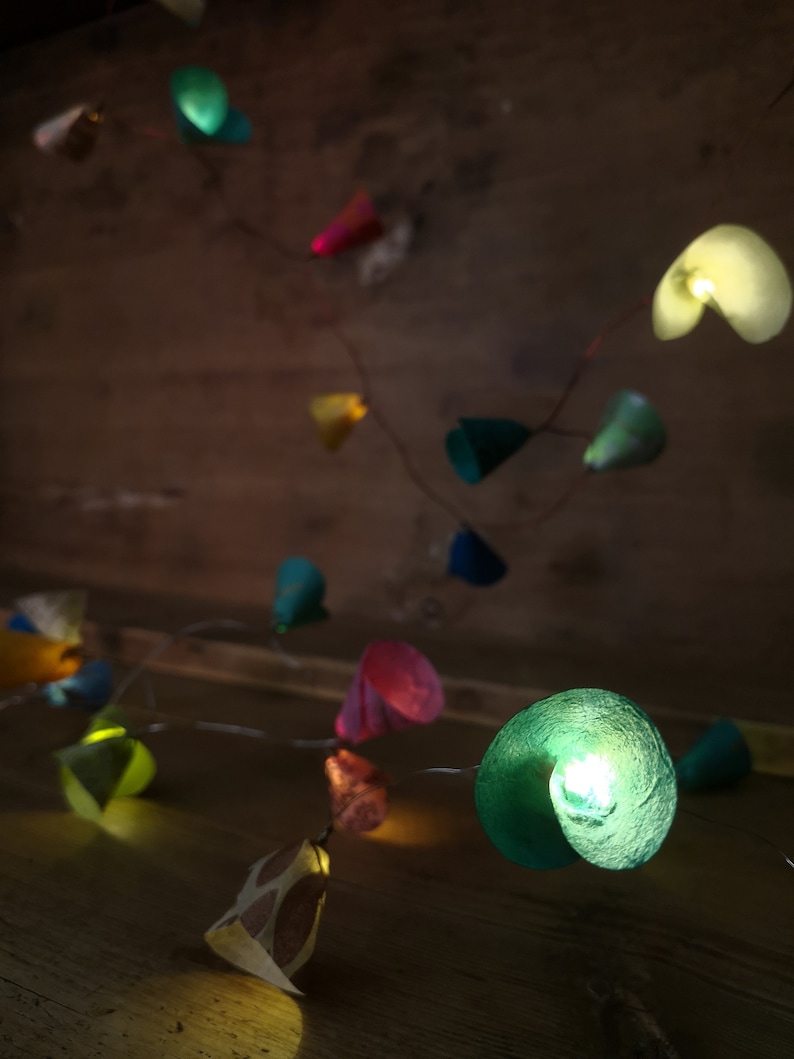 Puede incluir: Una cadena de luces de papel de colores con bombillas LED blanco c&aacute;lido. Las luces tienen forma de conos y c&iacute;rculos y est&aacute;n enhebradas en un alambre de cobre. Las luces cuelgan de una superficie de madera.