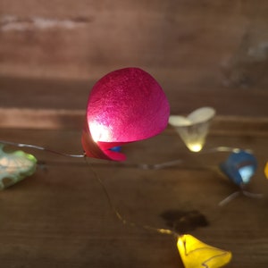 Puede incluir: Una cadena de luces con pantallas de papel de colores. Las pantallas tienen forma de coraz&oacute;n y est&aacute;n iluminadas. Las luces est&aacute;n enhebradas en un cable y cuelgan de una superficie de madera.