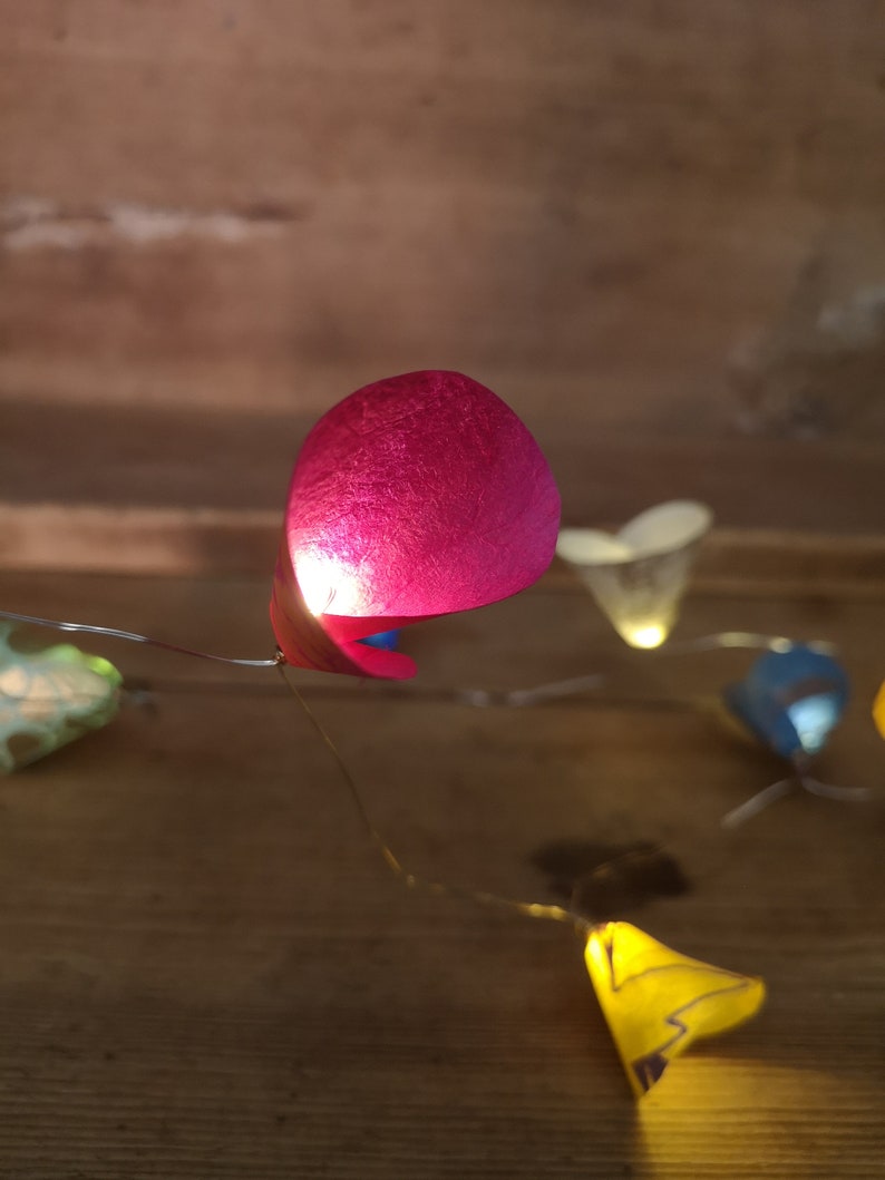 Puede incluir: Una cadena de luces con pantallas de papel de colores. Las pantallas tienen forma de coraz&oacute;n y est&aacute;n iluminadas. Las luces est&aacute;n enhebradas en un cable y cuelgan de una superficie de madera.