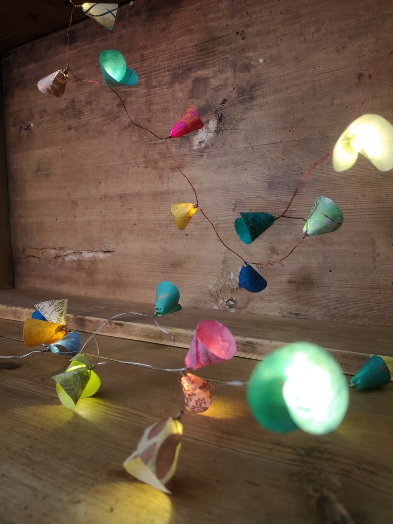 Puede incluir: Una cadena de luces con coloridos papeles en forma de cono. Las luces est&aacute;n enhebradas en un alambre de cobre y est&aacute;n encendidas, proyectando un brillo c&aacute;lido. Los papeles son de varios colores, incluyendo verde, azul, rosa, amarillo y naranja.