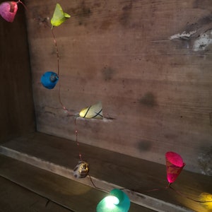 Puede incluir: Una cadena de luces con farolillos de papel de colores. Los farolillos tienen forma de flores y p&aacute;jaros. Las luces est&aacute;n enhebradas en un alambre de cobre y est&aacute;n encendidas. Las luces est&aacute;n colgadas en una superficie de madera.