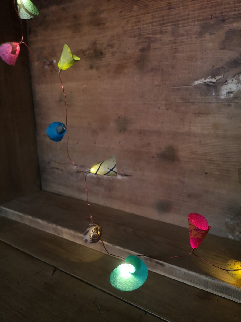 Puede incluir: Una cadena de luces con farolillos de papel de colores. Los farolillos tienen forma de flores y p&aacute;jaros. Las luces est&aacute;n enhebradas en un alambre de cobre y est&aacute;n encendidas. Las luces est&aacute;n colgadas en una superficie de madera.