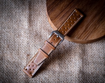 Puede incluir: Una correa de reloj de cuero marrón con costuras blancas y un diseño perforado. La correa tiene una hebilla plateada.