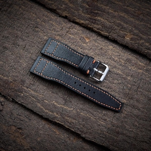 May include: Black leather watch straps with orange stitching and a silver buckle. The straps are made of textured leather and are displayed on a dark wooden surface. The stitching adds a contrasting detail.