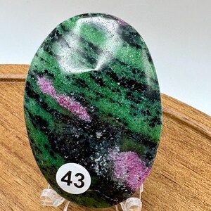 May include: An oval-shaped polished zoisite stone featuring vibrant green and deep black mineral inclusions with distinct patches of bright magenta ruby. A small white circular sticker with the black printed number 43 is affixed to the lower left side of the stone.