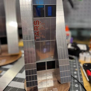 Può includere: Un modello argentato di un robot con la parola "TARS" stampata sul lato. Il modello è su una base di legno con il testo "TARS U.S. MARINE CORPS TACTICAL ROBOT" stampato su di essa.