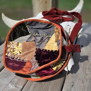 Puede incluir: Un bolso bandolera redondo confeccionado con un patchwork de diversas texturas y colores de tela, incluyendo terciopelo, pana y textiles estampados. El bolso presenta un borde naranja, una correa de pana de color rojo oscuro y costuras decorativas visibles.