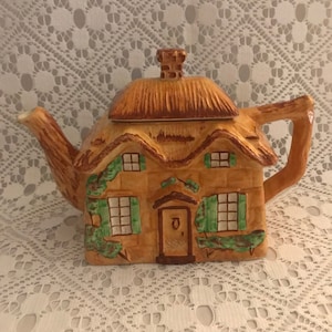 May include: A brown and green ceramic teapot shaped like a cottage with a thatched roof and green shutters. The teapot has a chimney and a door with a letter 'O' on it.
