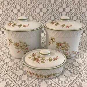 May include: Set of three white ceramic canisters with green trim and floral designs. The canisters have lids and are oval shaped.