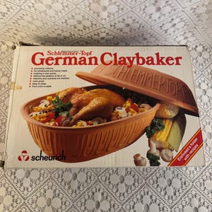 May include: A rectangular cardboard box for a Schlemmer-Topf German Claybaker. The box features a graphic of a terracotta-coloured clay baker filled with a roasted chicken, corn, and mushrooms, with a matching lid tilted to the side.