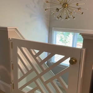 May include: White wooden baby gate with a geometric design, featuring a brass knob and hinges. The gate is installed at the top of a staircase and is open.
