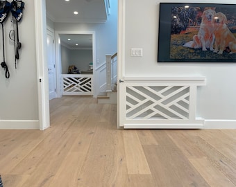 May include: White wooden pet gate with a geometric design, installed at the top of a staircase. The gate is open and there is a hallway with hardwood floors behind it. A picture of two golden retrievers is hanging on the wall to the right of the gate.