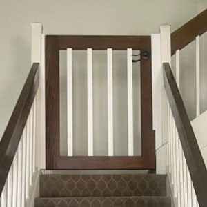 May include: A white wooden baby gate with a brown wooden frame is installed at the top of a carpeted staircase.