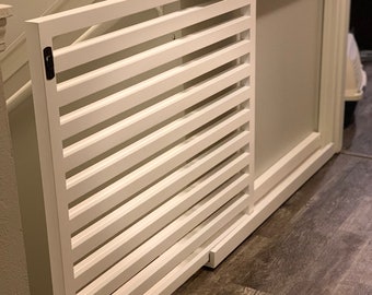 Op de afbeelding: Een witte houten traphek met horizontale spijlen, gemonteerd op een trap. De poort is gedeeltelijk open en toont het interieur. Het heeft een zwarte sluiting en kleine wielen voor gemakkelijke verplaatsing. De muren en vloer zijn neutraal van kleur.