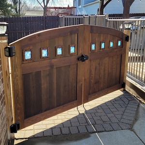 May include: A wooden double gate with an arched top and stained brown. The gate has a decorative design with blue glass panels. The gate is open and the background shows a brick patio and a wooden fence.