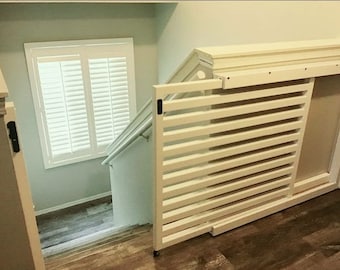 May include: A white wooden baby gate with horizontal slats is installed at the top of a staircase. The gate is partially open, revealing the steps and a window with white shutters. The walls are painted a light green color, and the floor is wood.