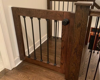 May include: A dark brown wooden baby gate with black metal spindles, a black knob, and a white trim. The gate is installed at the top of a staircase.