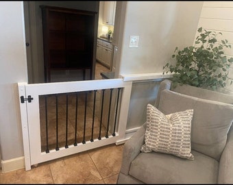 May include: White wooden gate with black metal bars, attached to a white wall. The gate is open and there is a gray armchair with a patterned pillow in the background.