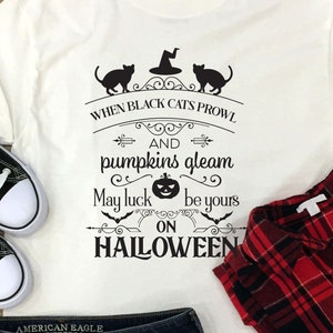 Puede incluir: Camiseta blanca con un gráfico negro que dice "When black cats prowl and pumpkins gleam, may luck be yours on Halloween". El gráfico presenta gatos negros, un sombrero de bruja, una calabaza y murciélagos. La camiseta está colocada sobre una superficie de madera con un par de zapatillas negras y blancas, un par de pantalones cortos de mezclilla y una camisa de cuadros rojos y negros.