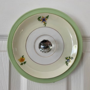 May include: A green and white ceramic plate with a floral design and a crystal knob in the center.